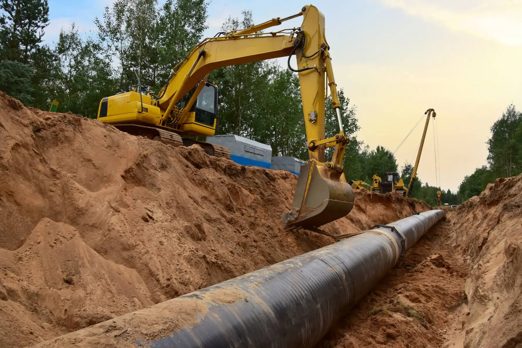 An excavator above an open trench with a large black pipeline laid along the bottom